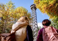 Zwei Personen mit Rucksäcken blicken auf einen hohen Aussichtsturm aus Metall, umgeben von herbstlich gefärbten Bäumen. | © Landratsamt Rhein-Neckar-Kreis, Sebastian Weindel