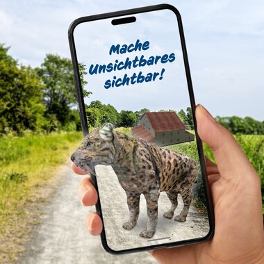 Hand hält Smartphone mit Darstellung eines Leoparden und Text 'Mache Unsichtbares sichtbar' vor ländlichem Weg mit Feldern und Bäumen im Hintergrund | © Landratsamt Rhein-Neckar-Kreis